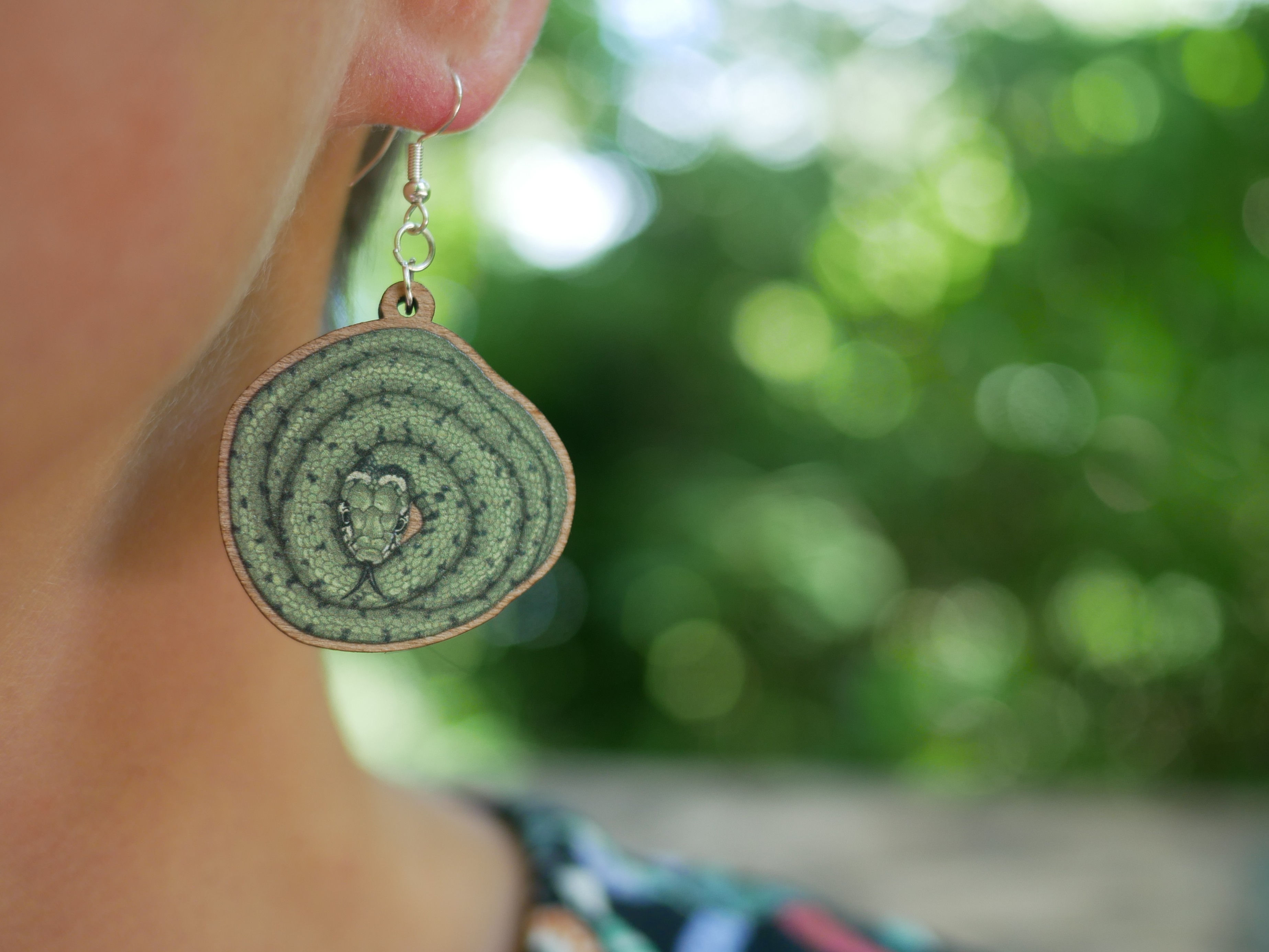 A wooden earring featuring an illustration of a grass snake, hung from an ear lobe