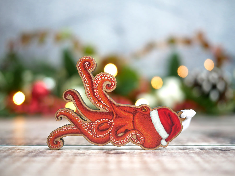 A wooden Christmas tree decoration of an illustrated octopus in a santa hat. On a sandy beach.