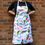 Thumbnail: White apron with illustrated parrots all over it. Worn by a model stood against a brick wall.