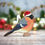 Thumbnail: Wooden Christmas tree decoration featuring an illustrated bullfinch wearing a santa hat. Ornament sat in front of lights