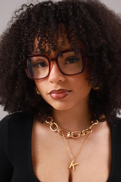 young woman with kinky curly hair looks at the camera. she wears a black shirt, a gold chain and big dark red glasses.