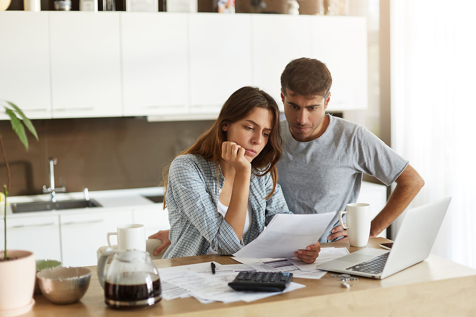 This photo shows a man and a woman standing in their kitchen, looking worried while studying some papers