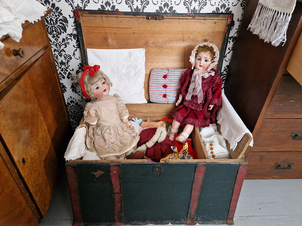 Antiikkinuket istuuvat kapioarkussa vanhojen nukenvaatteiden päällä. Talonpoikaistalon pikkukammarissa jossa taustalla mustavalkoinen isokuvioinen tapetti. Toisella antiikkinukella punainen samettimekko ja toisella beige mekko ja päässä rusetti.