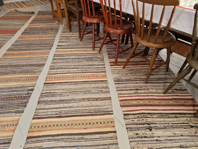 Harmaalla lankkulattialla talonpoikaistalossa on useita räsymattoja. Taustalla on puutuoleja pitkän pöydän ääressä. Väripaletti on maanläheinen ja rauhallinen.