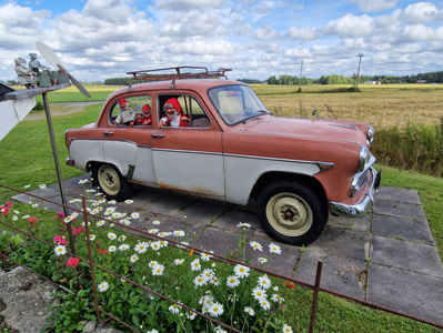 Vanha kaksivärinen veteraaniauto Mosse, maalaispihassa, kolme tonttua ikkunassa hymyilee, taustalla pelto ja pilvinen taivas.