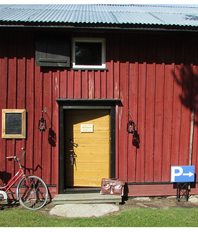 Punamullalla maalattu vanha makasiini jossa keltainen ovi joka on auki. Oven vieressä vanha polkupyörä ja salkku