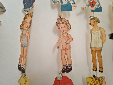 Kolme vintage-paperinukkea seinällä, yllään alusvaatteet. Yksi nukke vaaleanpunaisessa pitsiasussa. Tausta vaalea, neutraali. Nostalginen tunnelma