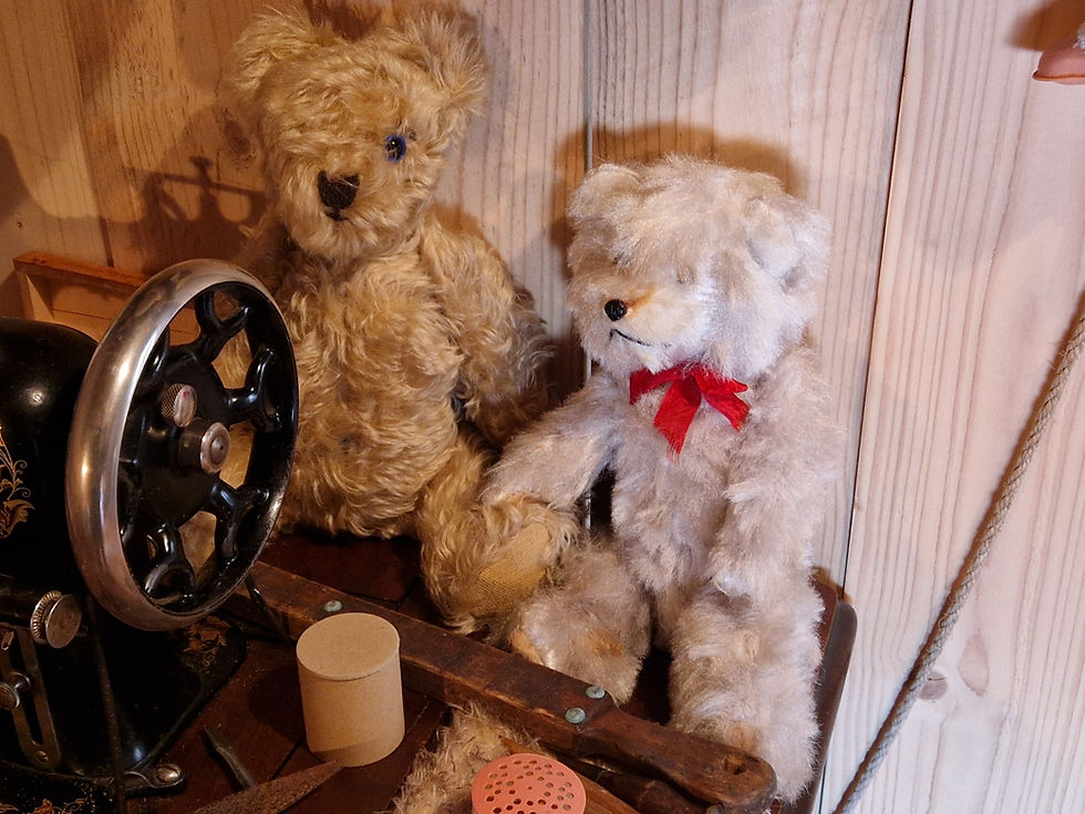 Kaksi pehmonallea istuu puista taustaa vasten. Vasemmalla vanha ompelukone, oikealla nalle punaisella rusetilla. Tunnelma on nostalginen.