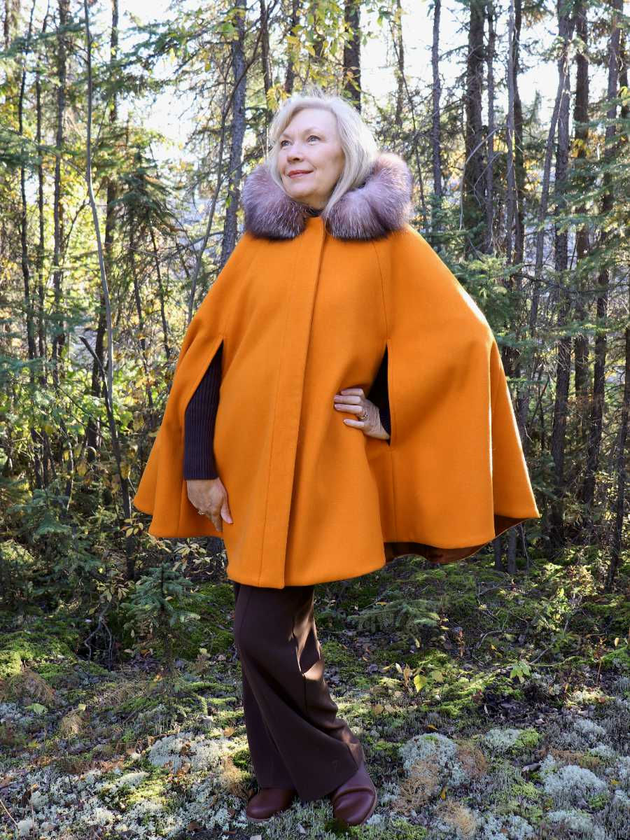 Thumbnail: In a forest, a woman wears an elegant, bright orange cape with brown fur trimming hood. The hood is down.