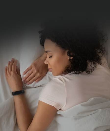 Woman peacefully sleeping in a bed, wearing a white shirt and a black fitness tracker. Soft lighting, white sheets, calm mood.