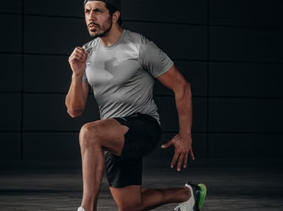 Man in a gray shirt and black shorts lunges forward on one knee, focused expression. Dark background, wearing white sneakers with green accents.