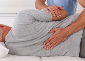 A person lies on their side in a gray shirt receiving a therapeutic back adjustment from a professional in a clinical setting.