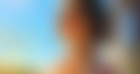 A young woman with curly brown hair, wearing a floral shirt, smiles while holding a glass of water and basking in bright morning sunlight on an outdoor patio with plants and a clear blue sky.