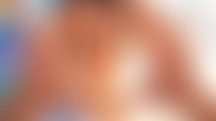 A woman with her hair in a bun framing her face with a heart shape made from her hands, standing in front of a blue wall covered in vision board photos.