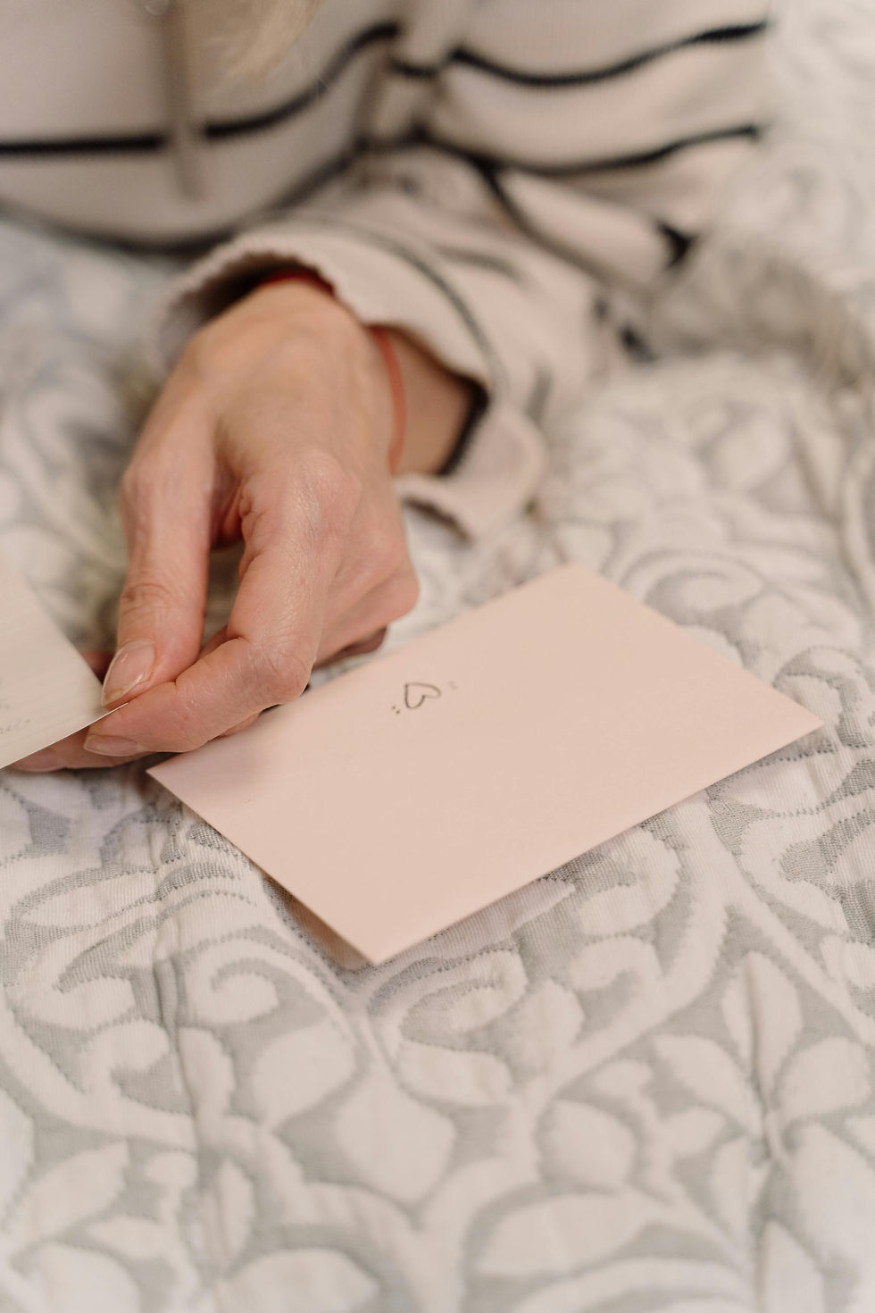 Hand holding a beige envelope with a heart drawing, resting on a patterned blanket. Person wears a striped sweater. Cozy, intimate mood.