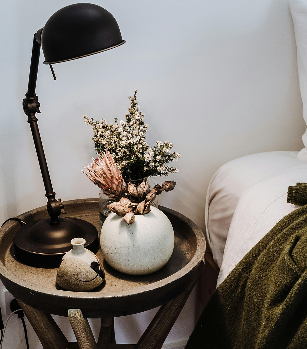 A round wooden table with a black lamp, white vase with flowers, and small pottery piece. Next to a white bed with a green blanket. Cozy setting.