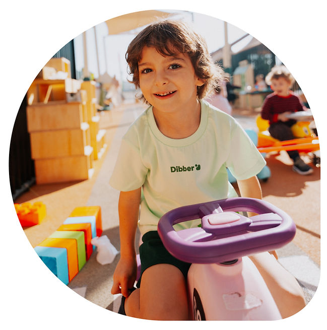 Child in a mint-green "Dibber" shirt smiles while riding a pink toy car outdoors. Bright blocks and another child playing in the background.