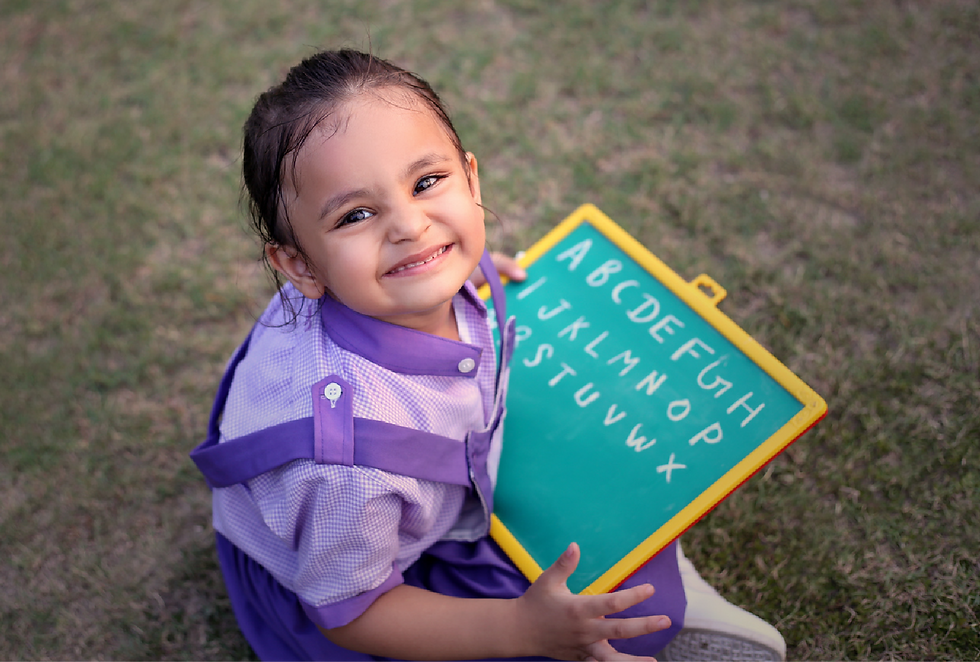 Smiling child in a purple outfit holds a small green chalkboard with the alphabet. Sitting on grass, cheerful mood.