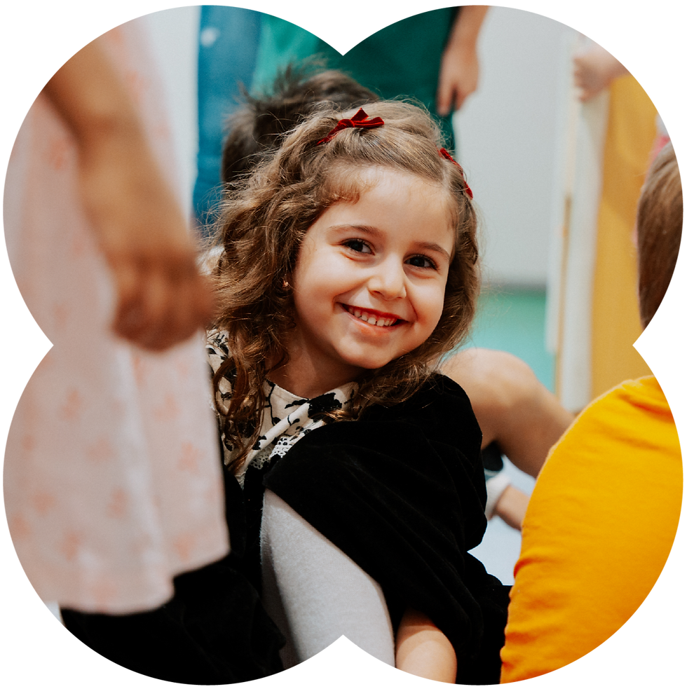 Smiling young girl with red hair bows among other children indoors, looking at the camera in a cheerful, playful scene.