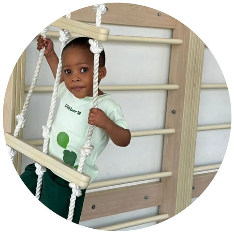 A child in a light green shirt climbs a rope ladder on a wooden play structure. The setting is indoors, with a calm and focused mood.