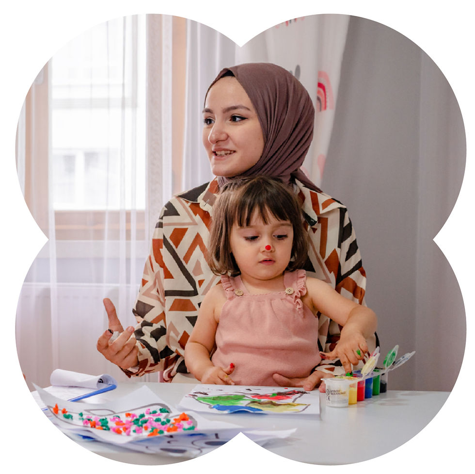 Woman in patterned top with young girl painting at a table. Bright paints and paper scattered. Light curtains in background, joyful mood.