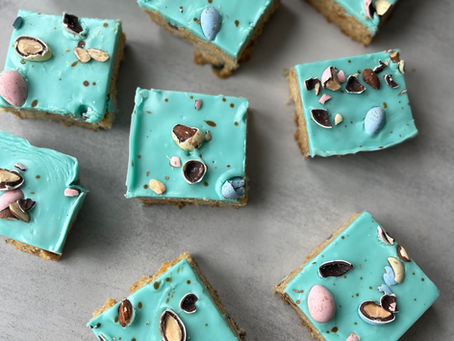 Square cookie bars topped with robin’s egg blue frosting, decorated with pastel candy eggs, chopped chocolate pieces, and cocoa speckles, arranged on a white plate and wooden tiered stand with Easter decor in the background.