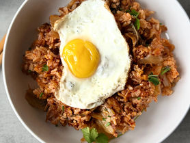 Skillet of kimchi ginger fried rice with visible pieces of kimchi, red onion, and ginger, lightly crisped, topped with a fried egg and served warm in natural light.