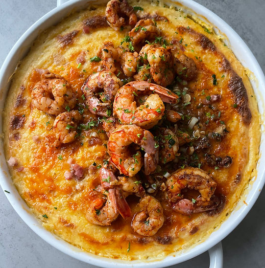 A large white deep dished filled with a creamy cheese grits casserole and topped with pancetta and prawns.