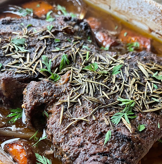 Close-up of a richly browned beef pot roast garnished with rosemary and fresh herbs, surrounded by carrots and onions in a rustic roasting dish.