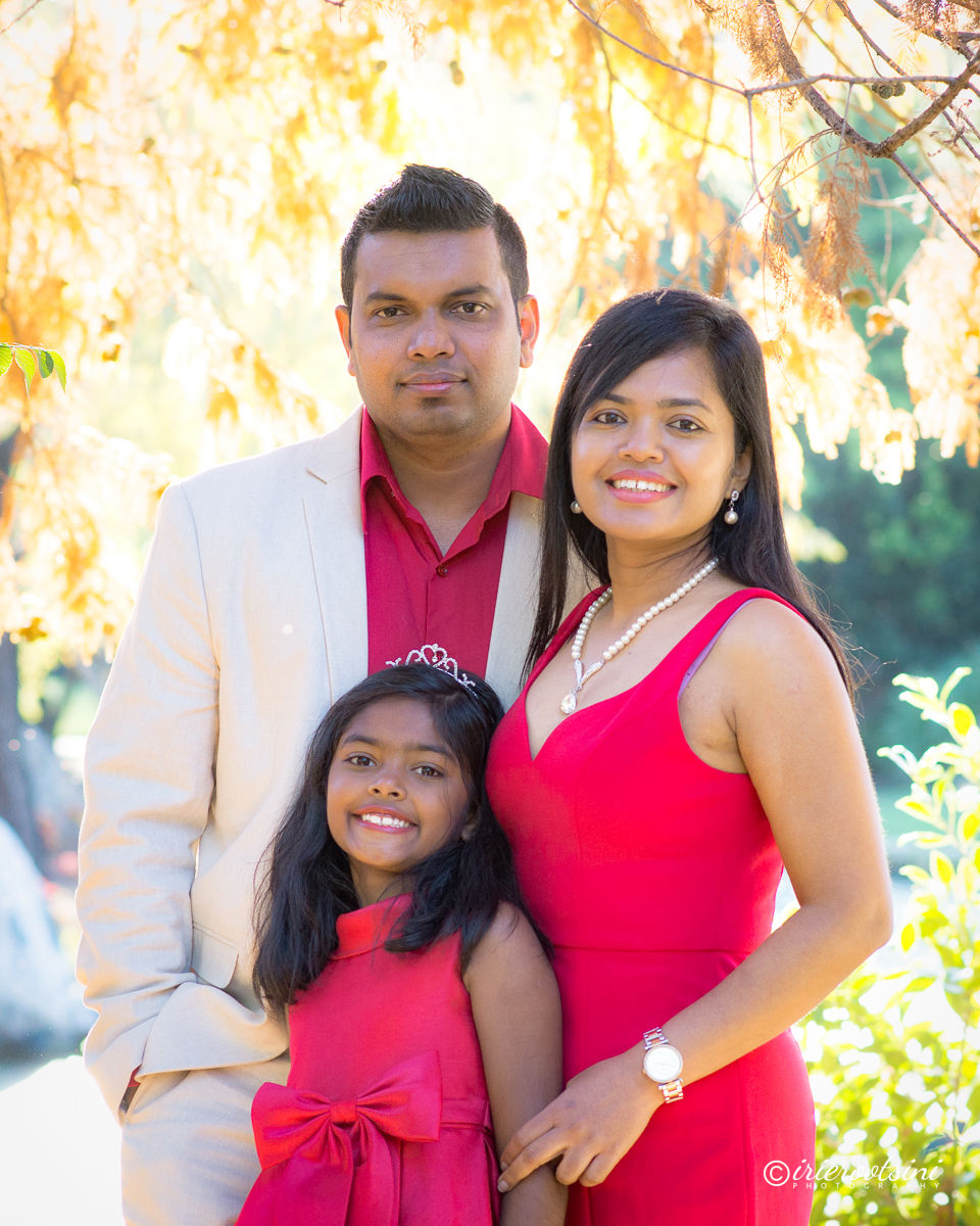 Family of three standing side by side in coordinated red outfits, smiling toward the camera in an outdoor garden.