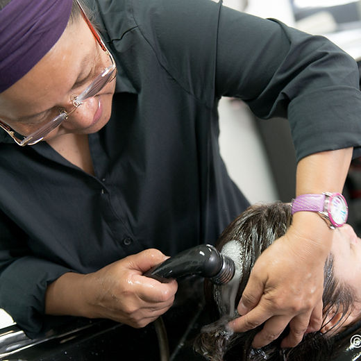 Client seated with head leaned back at wash basin while staff rinses her hair, showing suds from shampoo