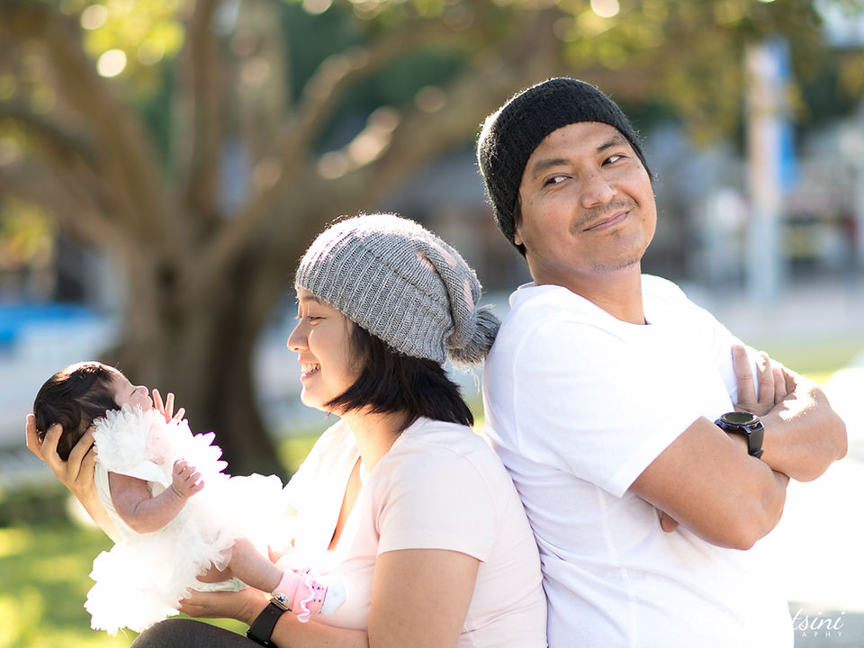 Parents sitting back-to-back holding their baby outdoors under soft morning light.