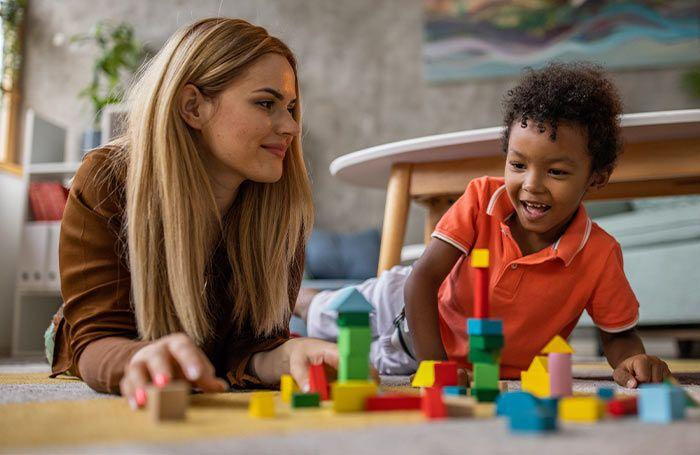 Nanny and young boy play on the floor with colorful blocks in a bright living room, smiling and focused together.