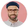 Smiling man with beard, glasses, and blue beanie on pink background.