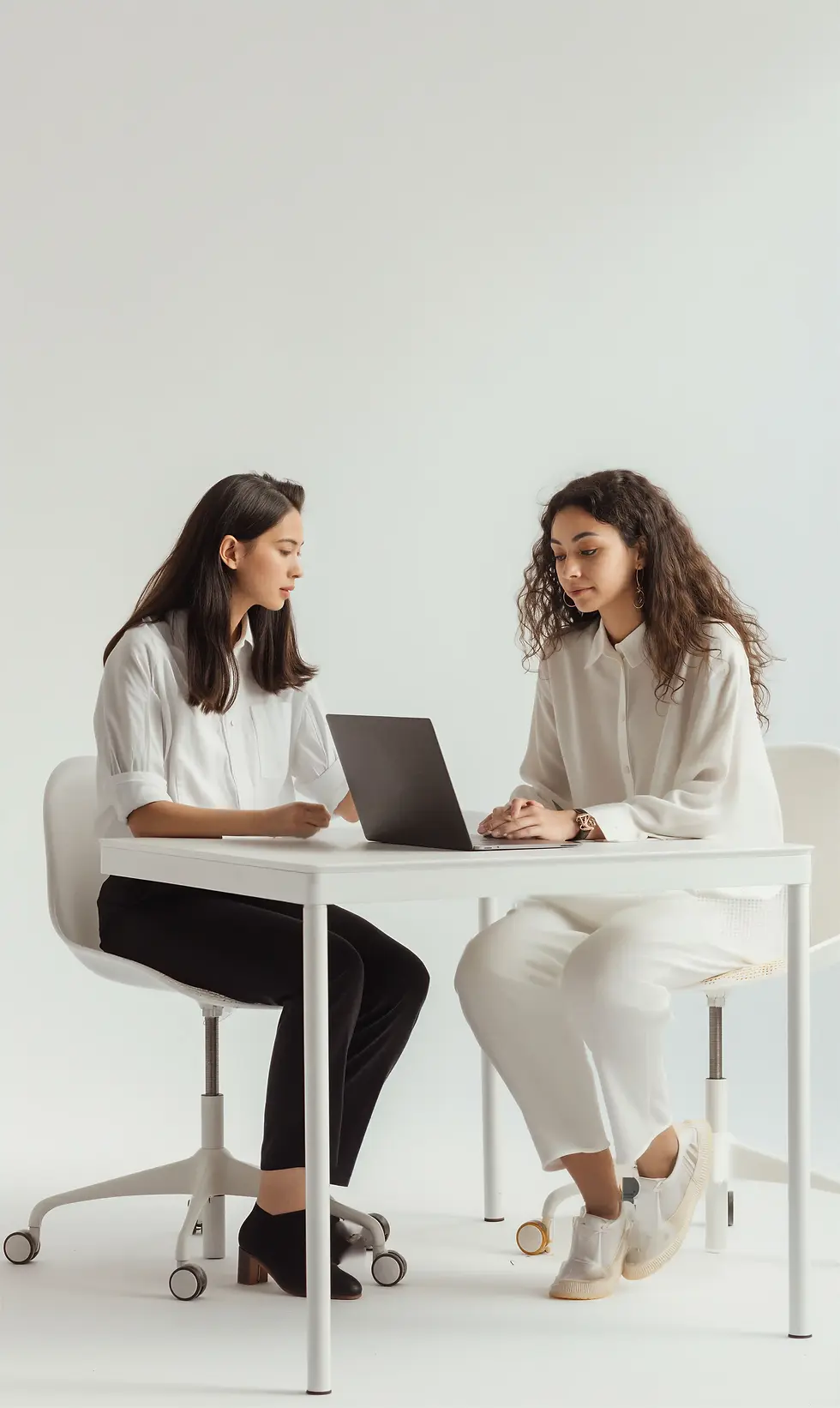 Two young women collaborating on a laptop at a white desk.