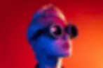 Person with short hair wearing steampunk goggles against dramatic red and blue lighting.