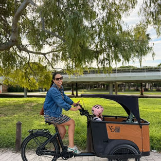 Family cargo bike ride in a green park in Valencia, with a smiling woman and a child in a pink helmet.