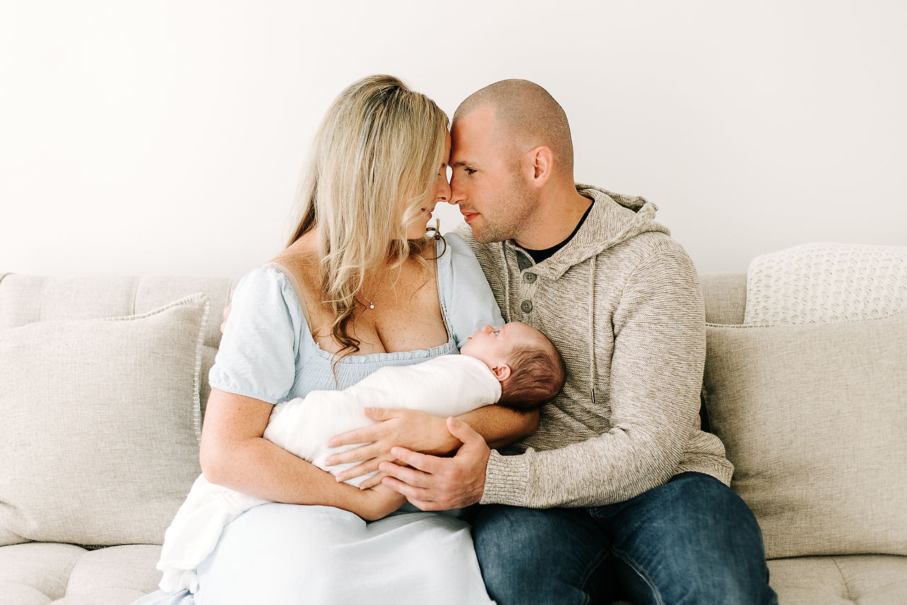 Family of three during a studio newborn session in Guelph, Ontario featuring coordinated outfits and a swaddled newborn baby on a couch