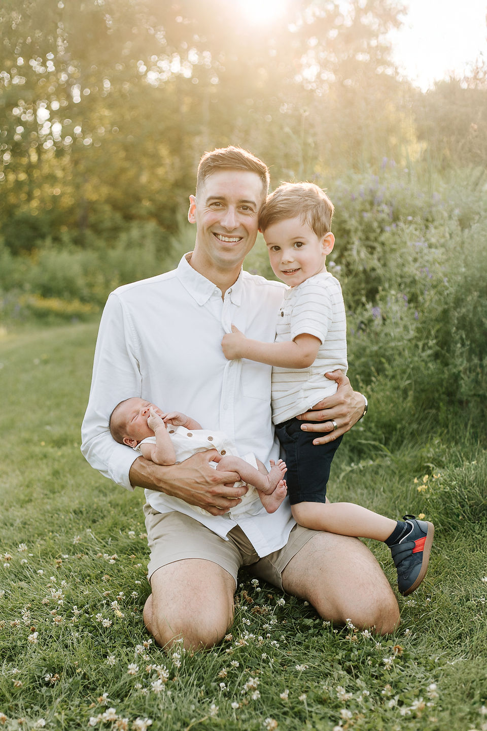 Outdoor newborn session at The Guelph Arboretum featuring a father holding his older son and newborn baby during golden hour