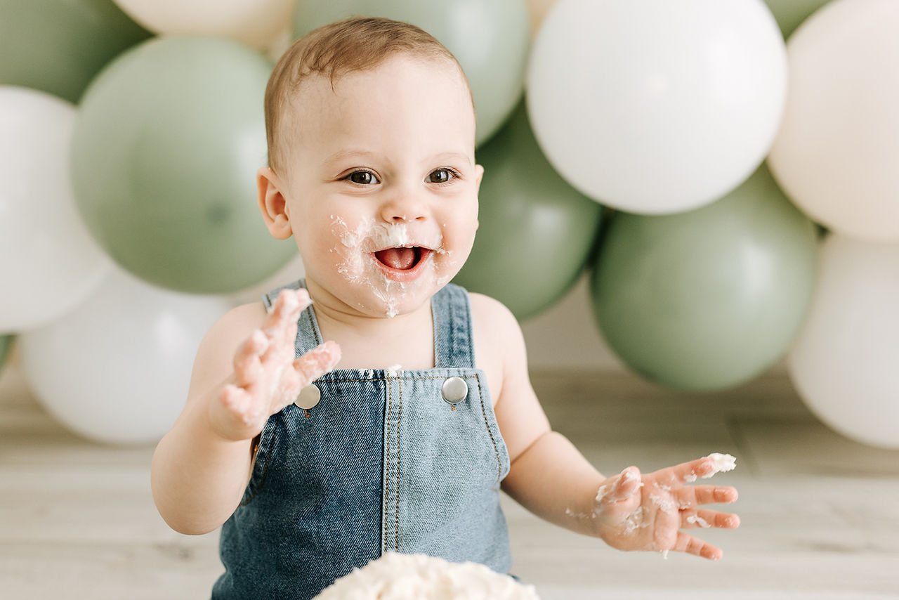 Baby in overalls with cake-covered hands during a From Maternity to Milestones cake smash session in Guelph, Ontario