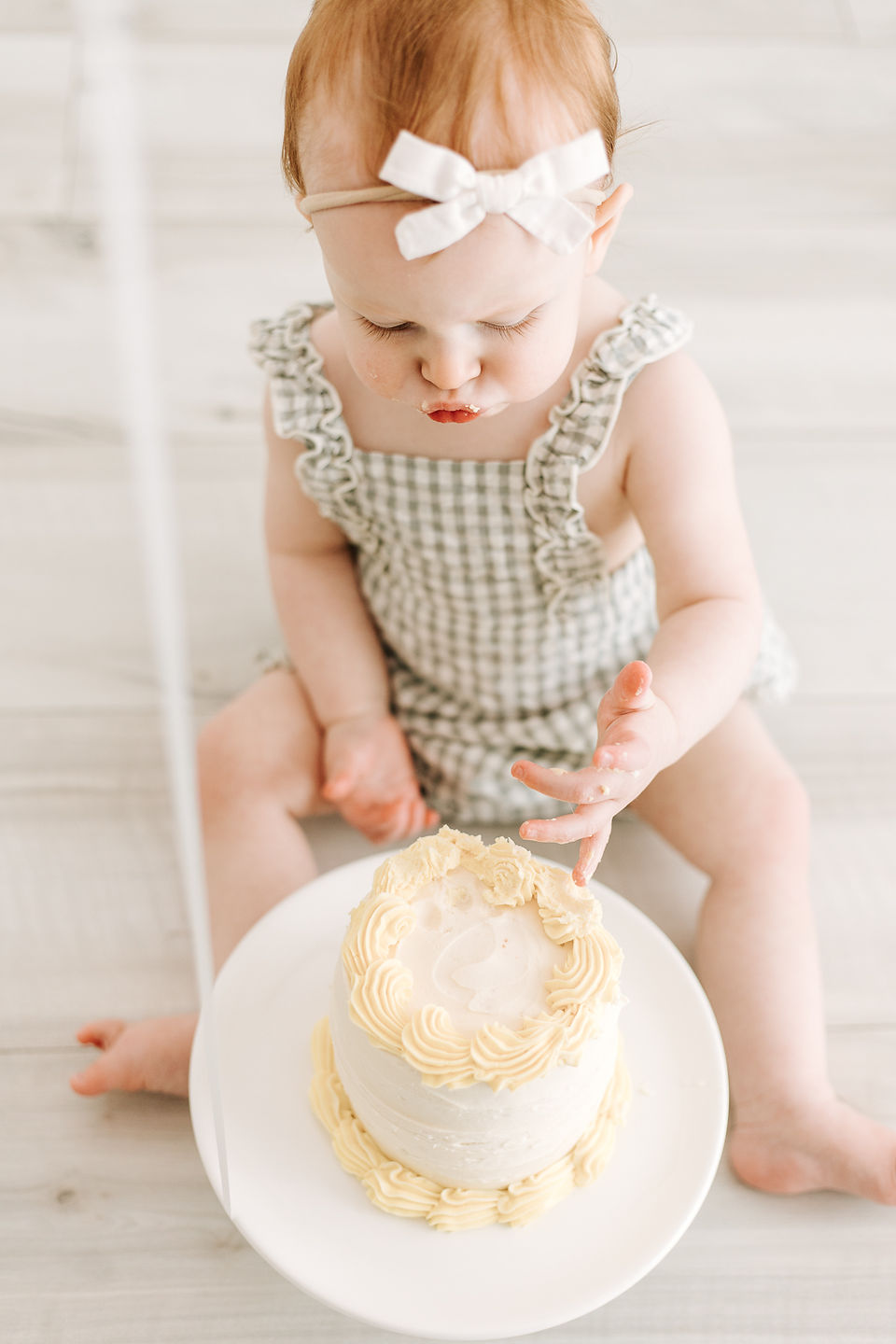 Baby girl sitting in front of a white cake during a cake smash session in Guelph, Ontario wearing a light patterned romper and bow headband