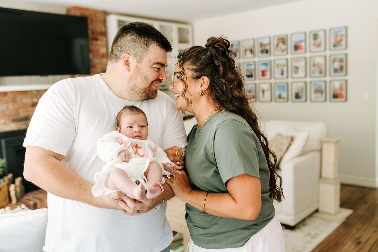 In-home newborn session in Guelph, Ontario featuring parents smiling together while dad holds their newborn baby in the living room