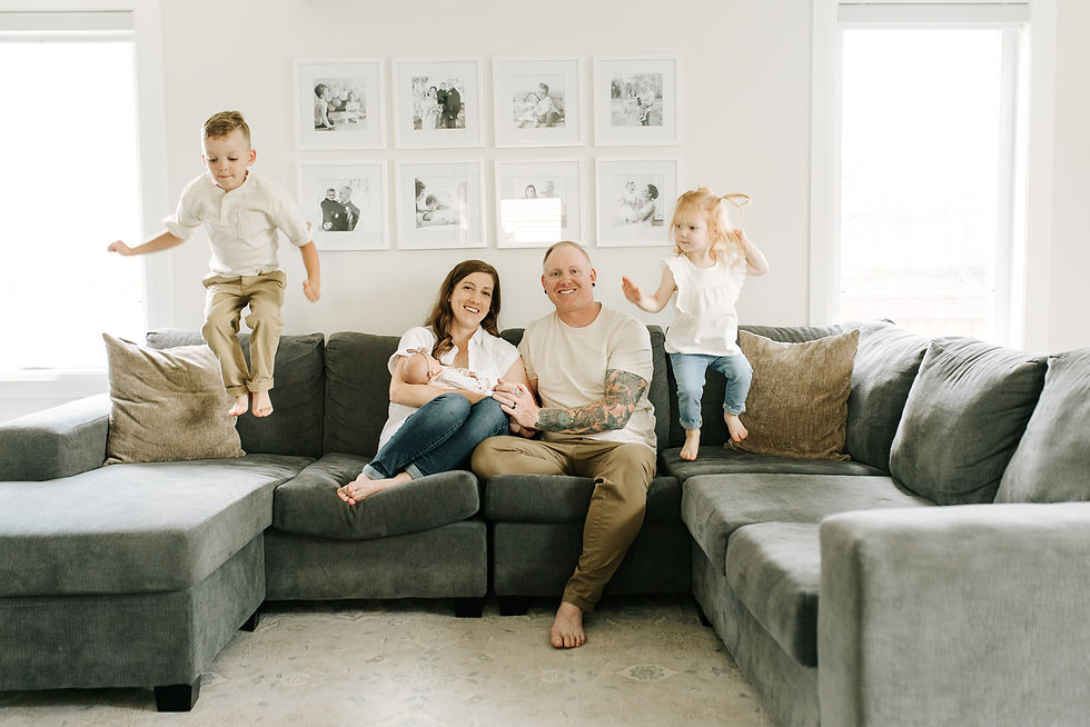 Family welcoming their newborn baby during an in-home newborn session in Guelph, Ontario, with two playful siblings jumping on the couch.