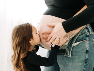 Maternity session photo of big sister-to-be kissing baby bump while dad embraces mom from behind in Guelph, Ontario