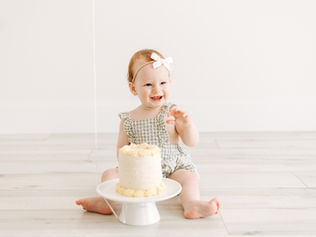 Smiling baby girl during a cake smash session in Guelph, Ontario wearing a gingham romper and sitting beside a small white cake