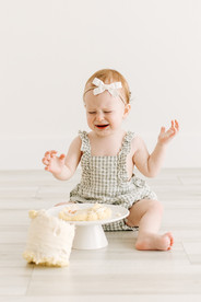 Baby girl sitting in front of a white cake during a cake smash session in Guelph, Ontario wearing a light patterned romper and bow headband