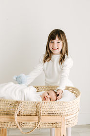Newborn session in Guelph, Ontario featuring big sister smiling beside newborn baby in bassinet in a clean, minimal studio setting