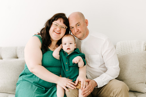 Family of three sitting on a couch during a milestone session in Guelph, Ontario with coordinated green outfits and a bright studio background