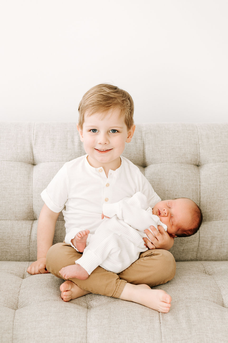 Big brother holding sleeping newborn baby during a newborn session in Guelph, Ontario on a light gray sofa with a clean studio background