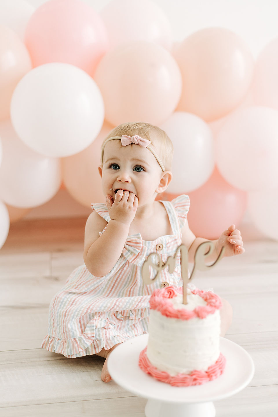 Baby girl enjoying her first birthday cake smash in Guelph, Ontario with a pastel balloon backdrop and pink and white birthday cake.